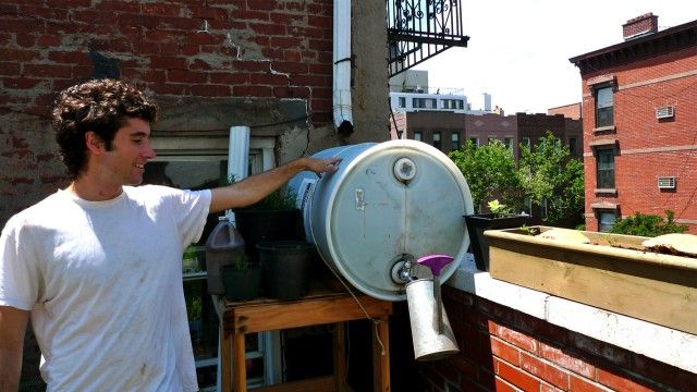 Patrick Kiley at the rainwater collector. Hay mulches on a taller section of rooftop, and rainwater washes through the mulch, traveling down the gutter into the collection barrel seen here. Cirell converted a food-safe detergent barrel he "borrowed" from his day job at the Greenpoint Beer Works to make the collector. It provides water for the garden. 